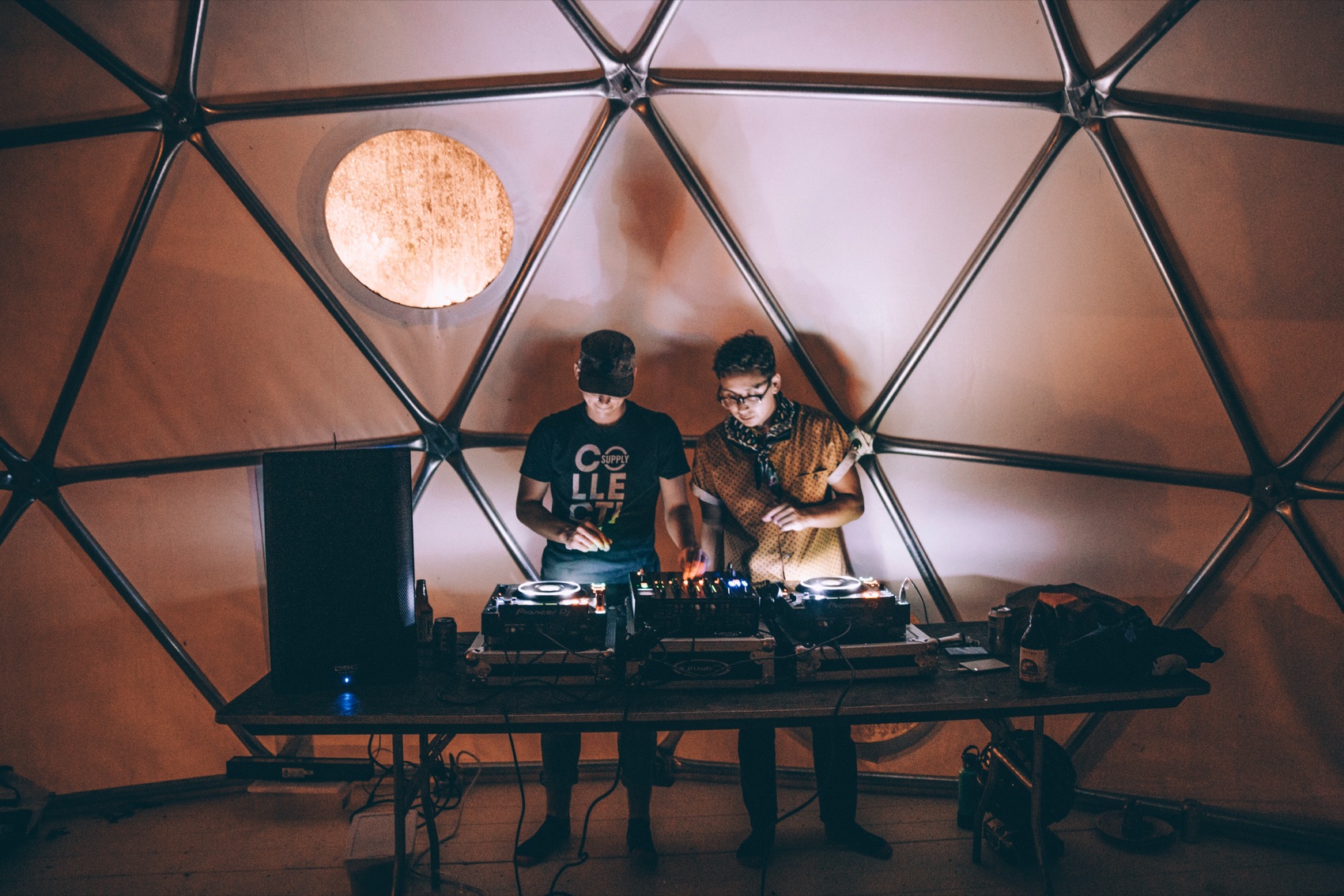 Two DJs, Andrew Smith at left in a Supply Collective shirt, playing inside a geodesic dome lit by one round window.